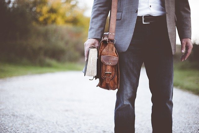 man with satchel bag on his right shoulder holding a book in his right hand down by his side